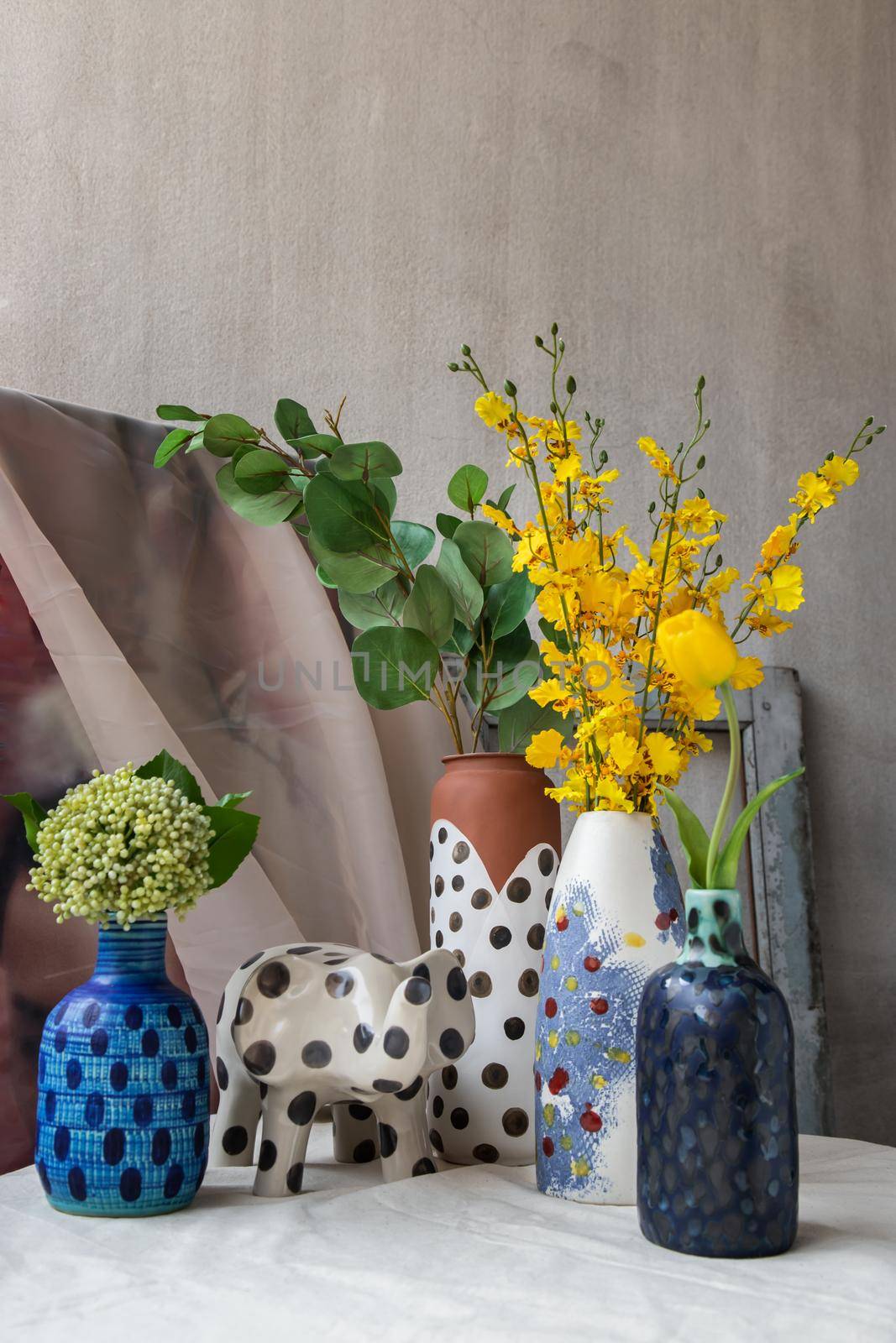Bangkok, Thailand - Feb 06, 2021: Bouquet of many flowers in Handmade Assorted Many Different Ceramic Polka Dotted Vases and Elephant shaped ceramic on white textured table cloth. Home decor, Space for text, Selective focus.