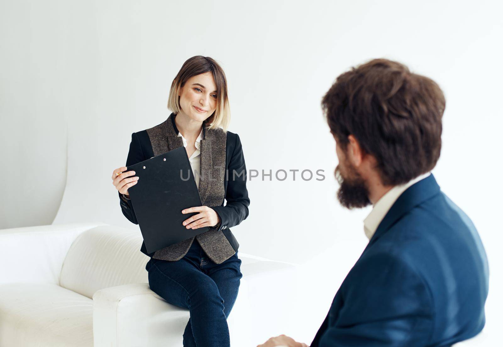 A man in a blue jacket sits opposite a woman in a suit on a light background indoors. High quality photo