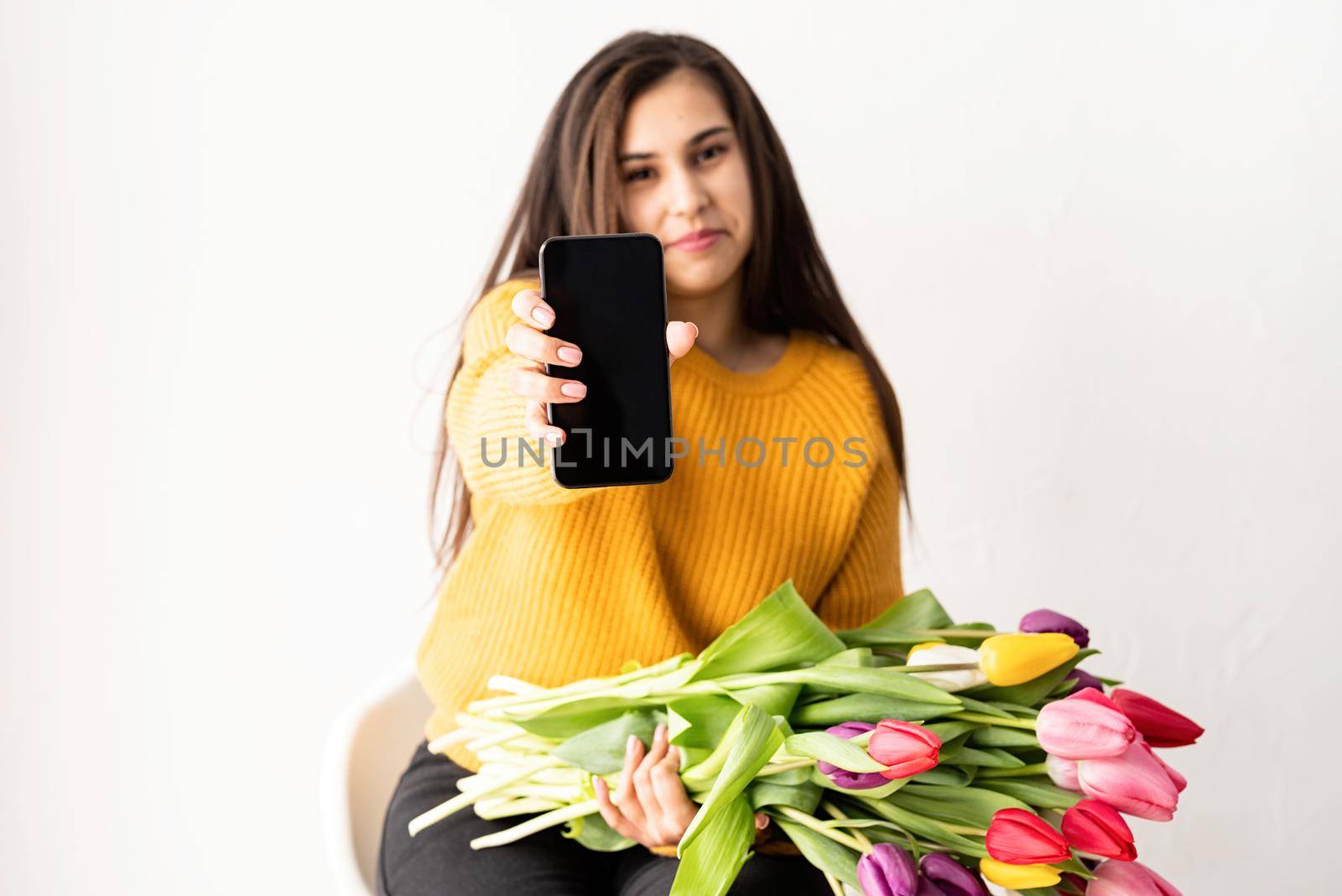 Beautiful young brunette woman in yellow sweater with bouquet of fresh pink tulips and blank mobile phone for mock up