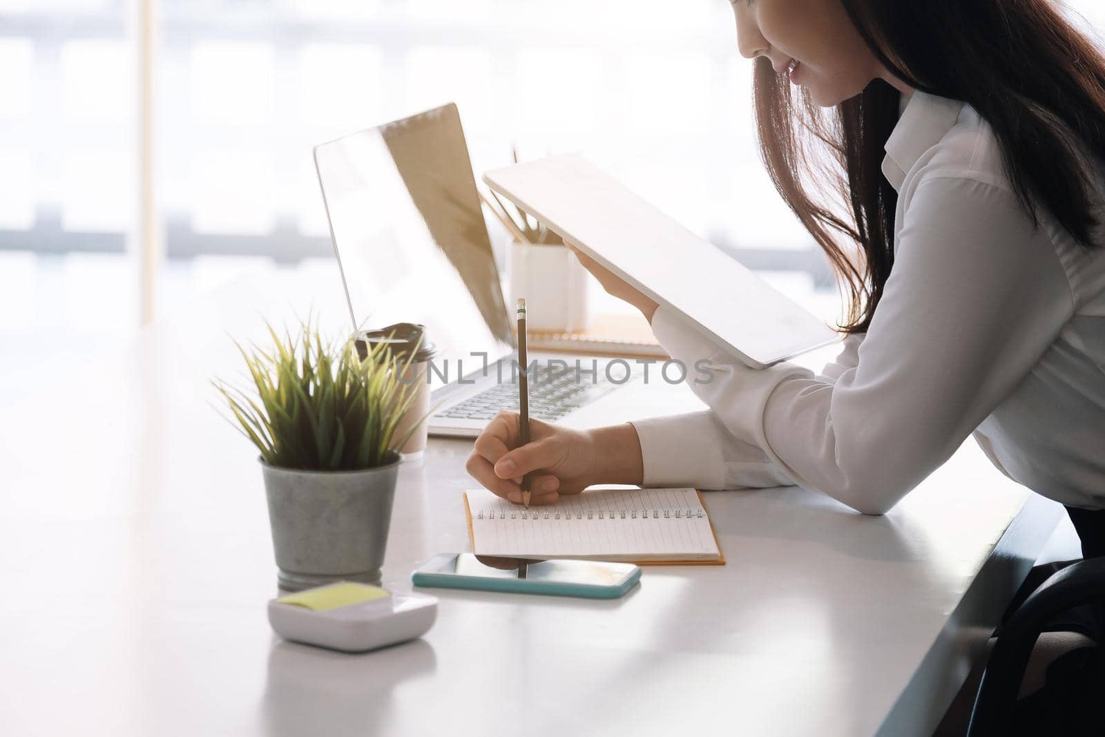 Photo of cheerful joyful asian woman writing down notes holding tablet for data analysis.