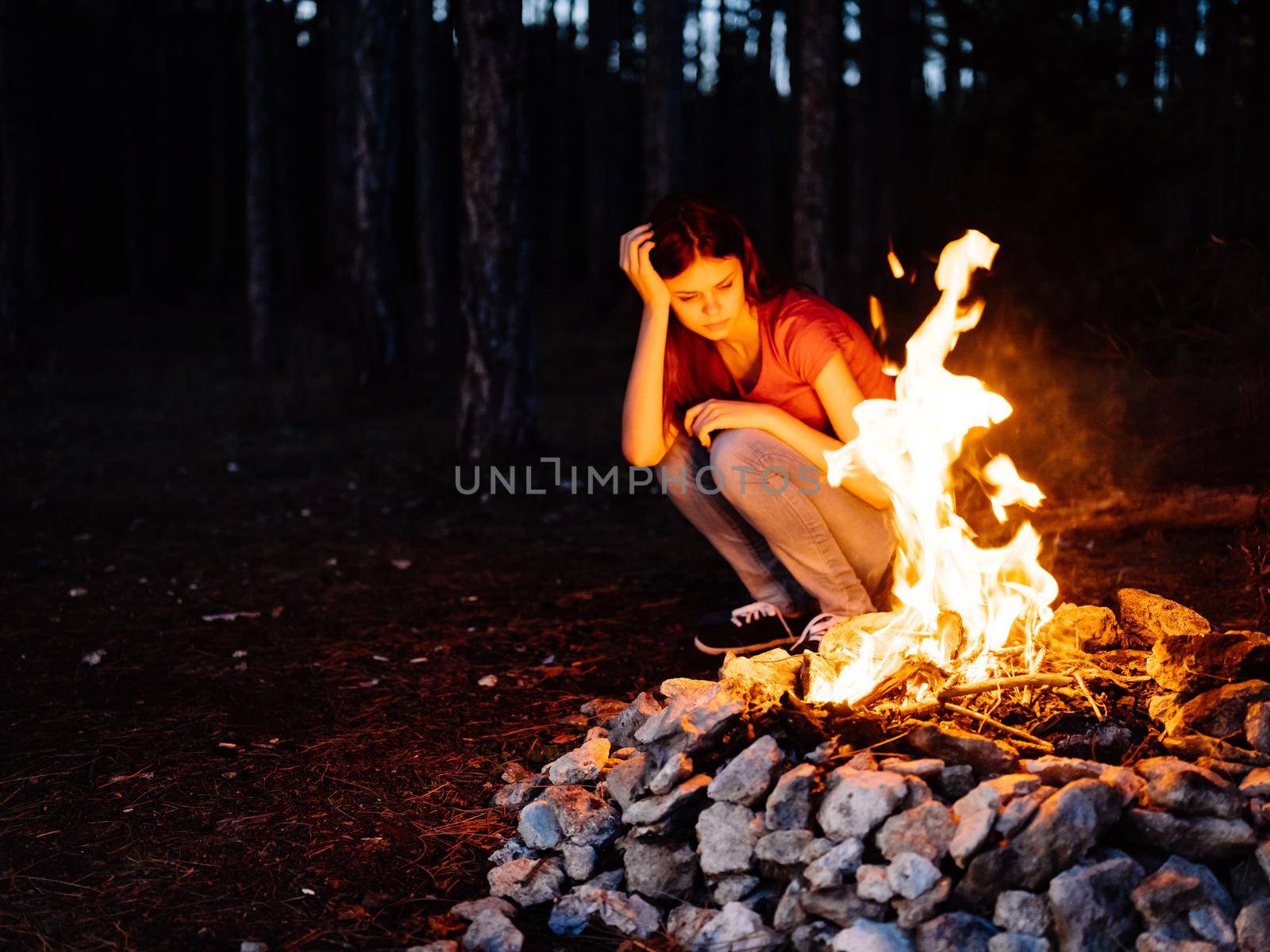 A woman sits near the fire and looks at the fire in a pine forest. High quality photo