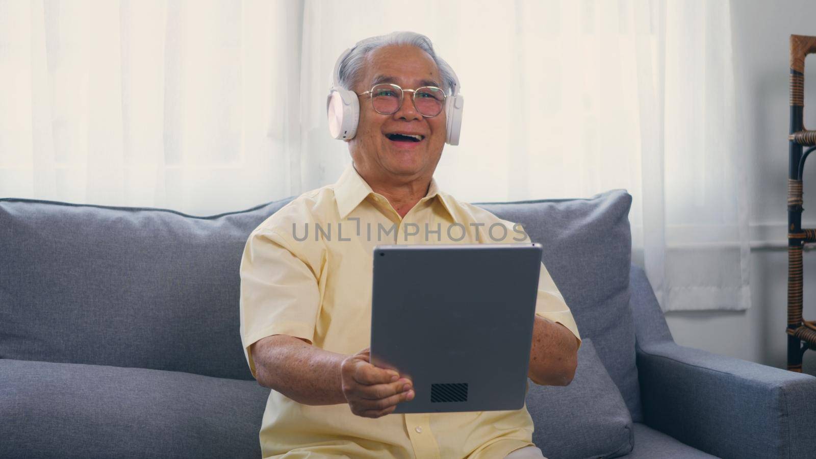 Old man grandfather smile with eyeglasses relaxing wear headphones is listening to music using a digital tablet, Asian happy senior old gray-haired sitting in living room on sofa at home video call