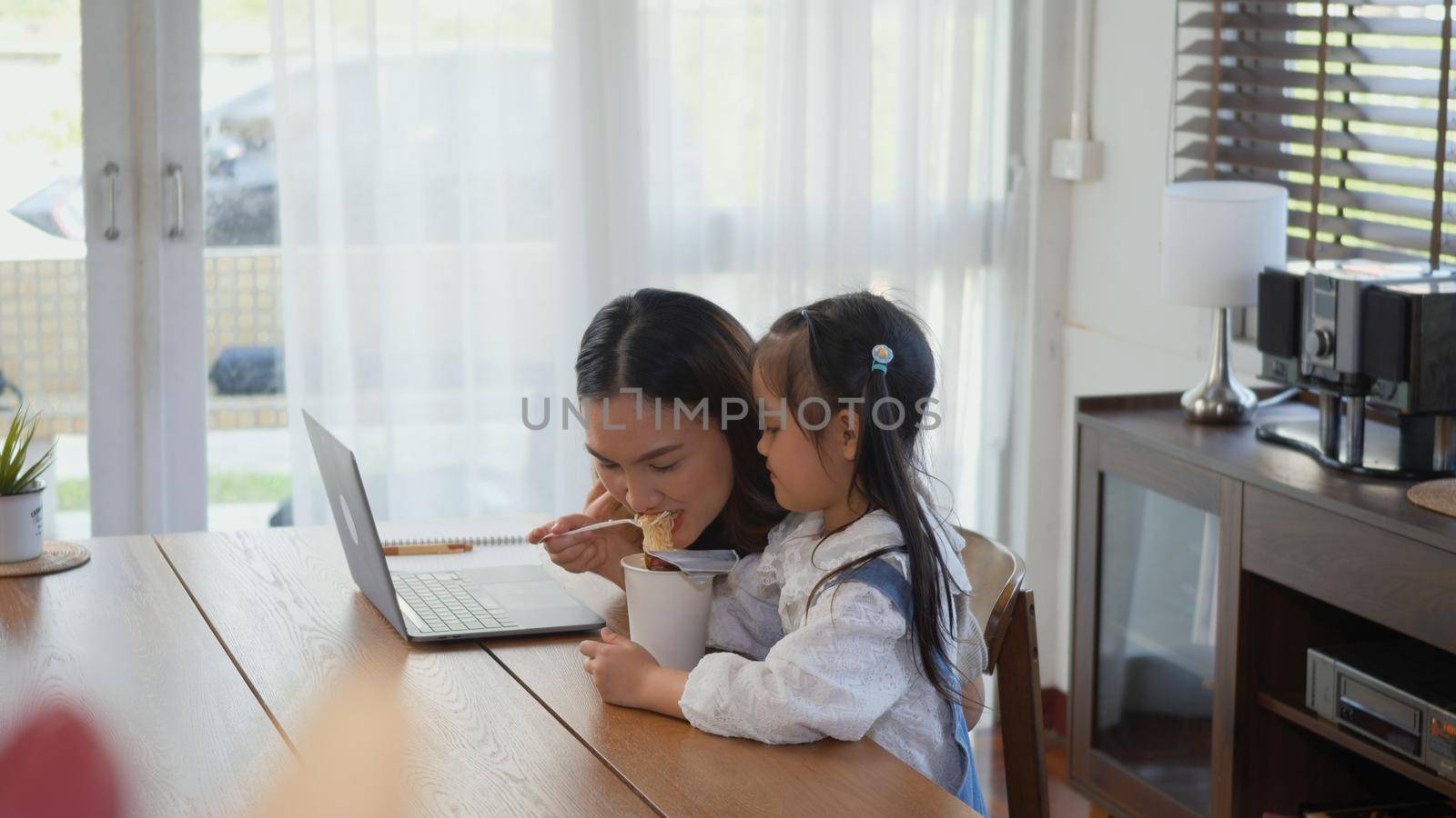 Asian family young mother working on laptop computer and her little daughter bring instant noodles to mom at home, woman eats noodles while doing work from home