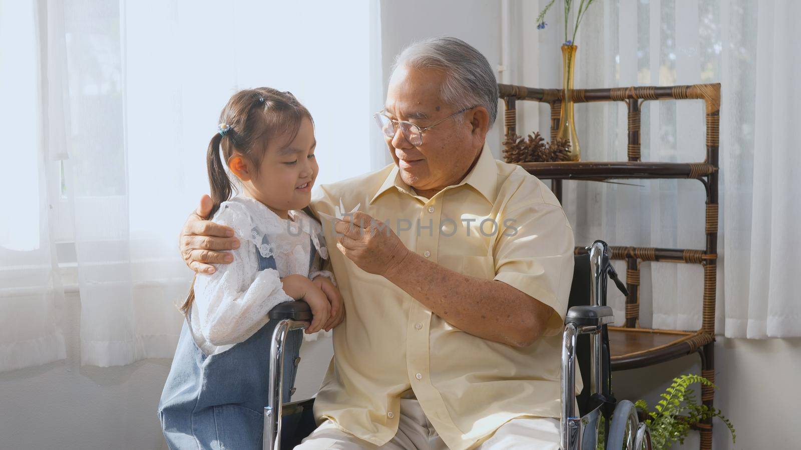 Asian elderly person sitting lonely in a wheelchair, sick and granddaughter running come holding a paper bird to play with and encourage, Happy female old senior man and little child