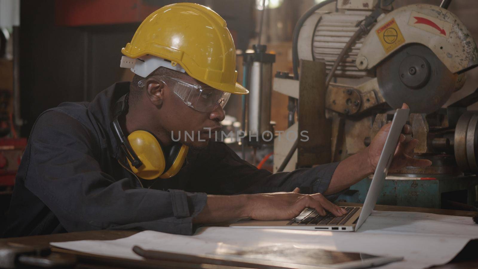 worker man with yellow helmet and ear protection typing keyboard of laptop computer by Sorapop