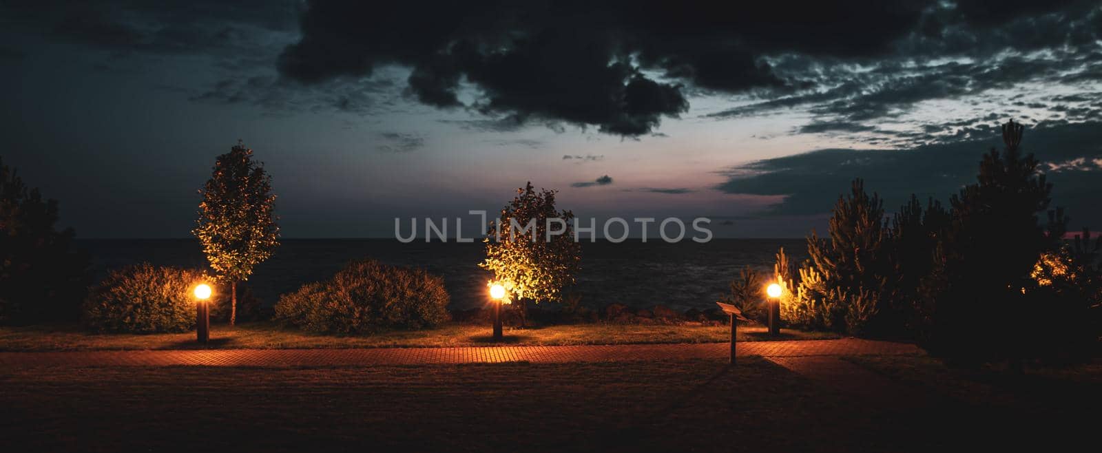 Evening on the seaside with a path lit by lanterns, bushes and pines and dramatic clouds