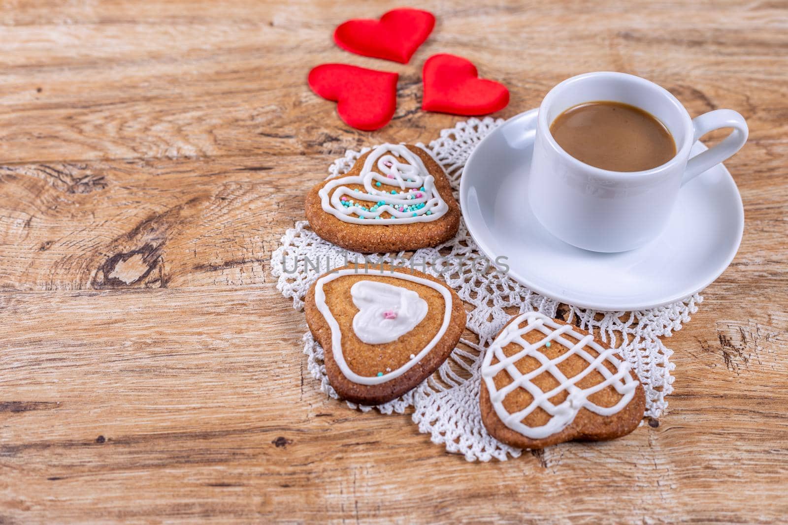Homemade heart-shaped cookies, decorated with white icing with decorative sprinkles, and red hearts on a wooden table with a cup of coffee, top and side view.