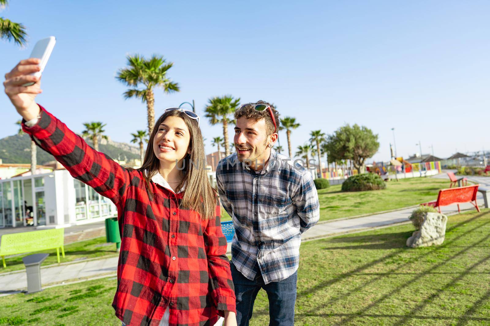 Young happy couple outdoor taking selfie portrait with smartphone smiling in a city park of sea ocean resort at sunset or dawn. Two friends having fun in the green using social network technology by robbyfontanesi