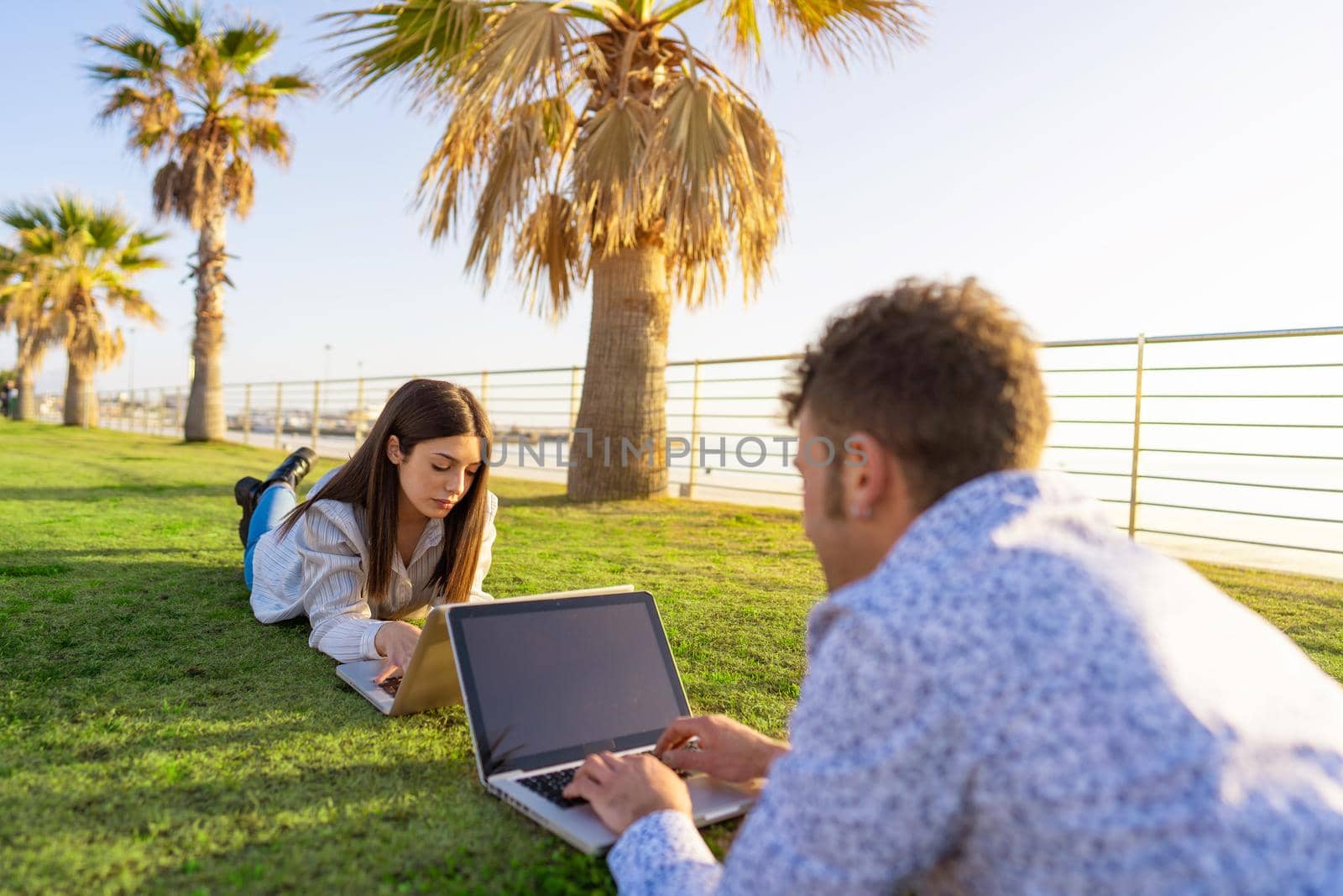Young couple lying on the grass in smart working with laptop facing each other at sunset or dawn. New normal activity of job or study outdoor with internet wireless connection technology