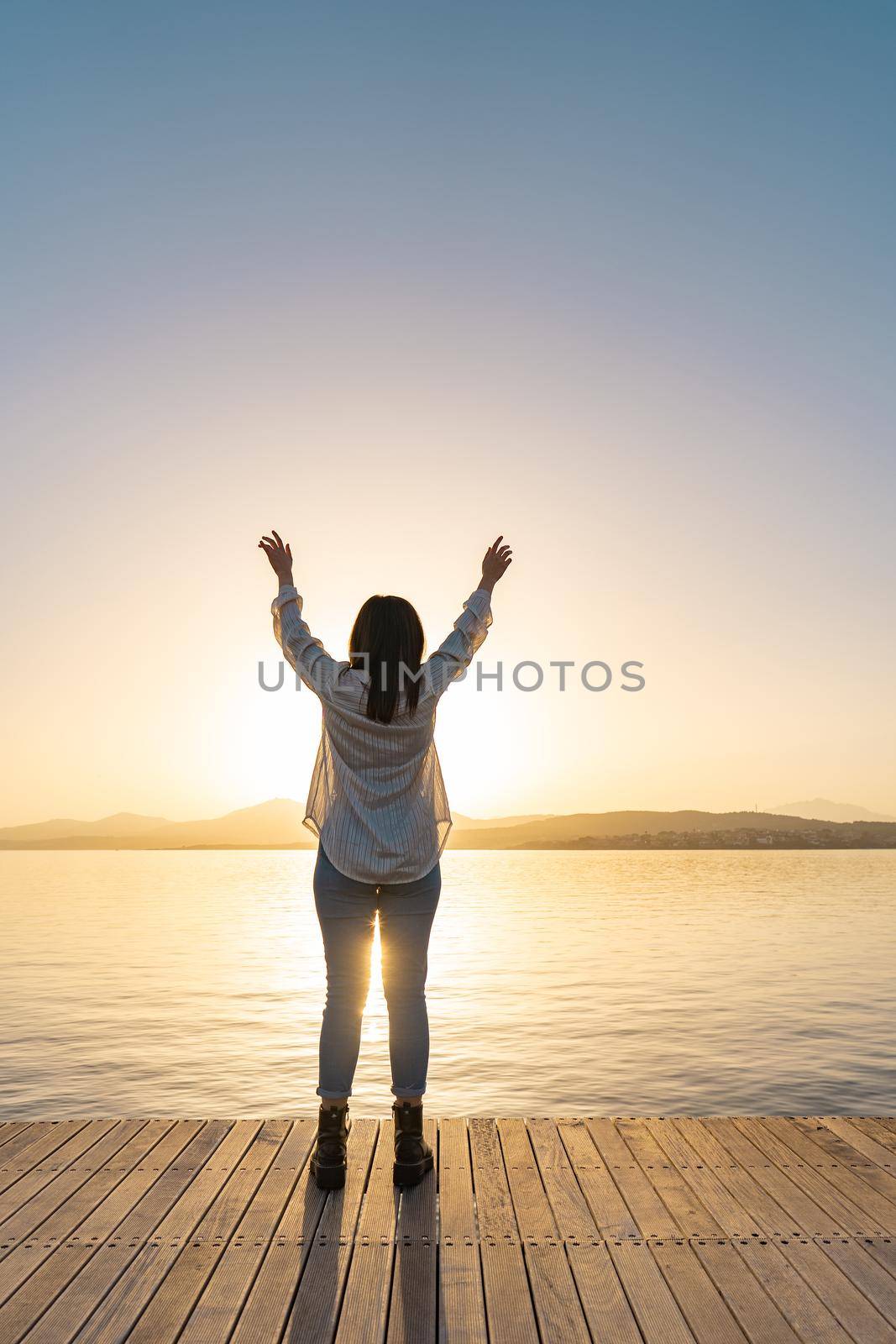 Young woman inspired by the sun open arms to the sky looking sea ocean water at sunset or dawn. Back view silhouette in the backlight of spiritual girl thanks God in religious gesture position