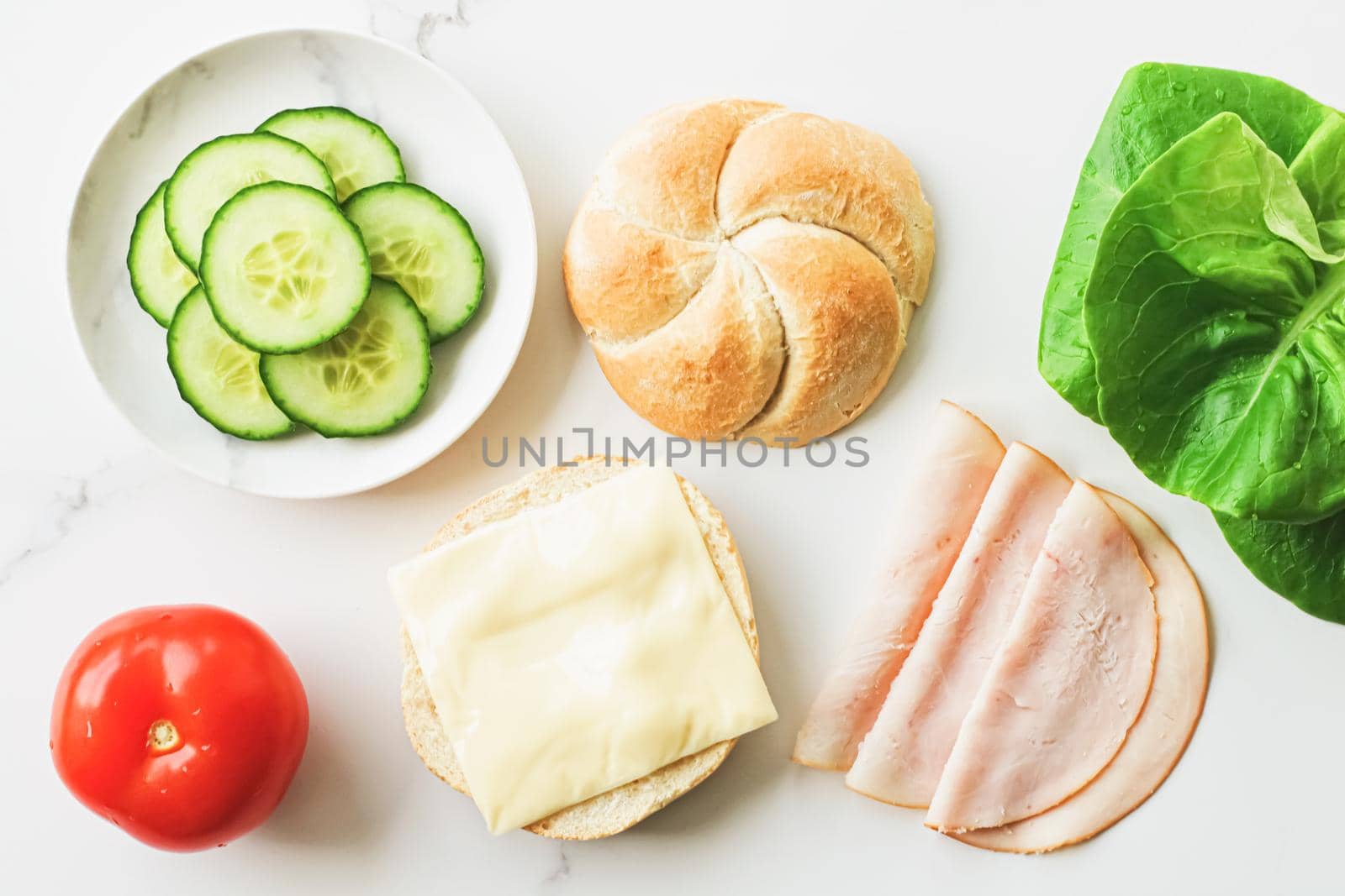 Food products and ingredients for making sandwich. Ham, cheese, burger bun, lettuce, cucumber and tomato as recipe flatlay on marble kitchen table background