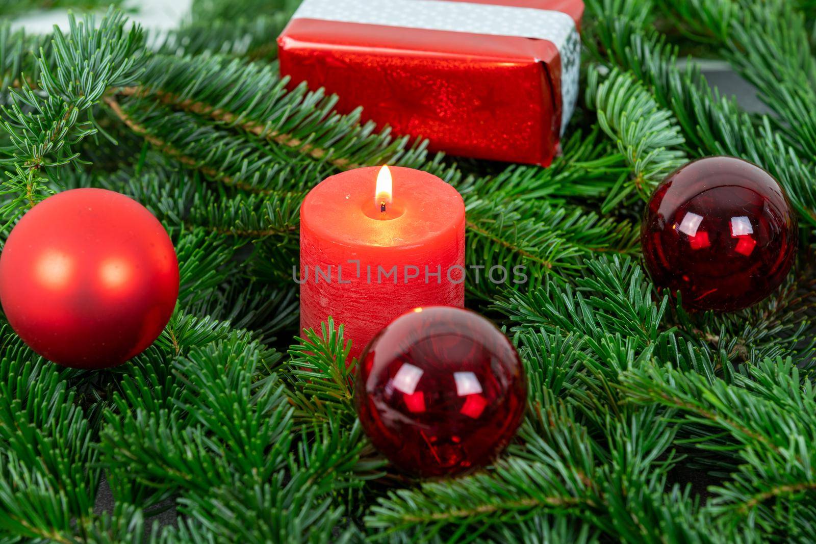 Atmospheric christmas motif with red burning candle surrounded by Nordmann fir branches, red christmas tree balls and gift parcel
