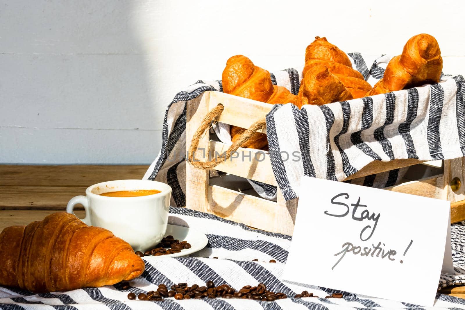 Coffee cup and buttered fresh French croissant on wooden crate. Food and breakfast concept. Morning message “stay positive” on white board