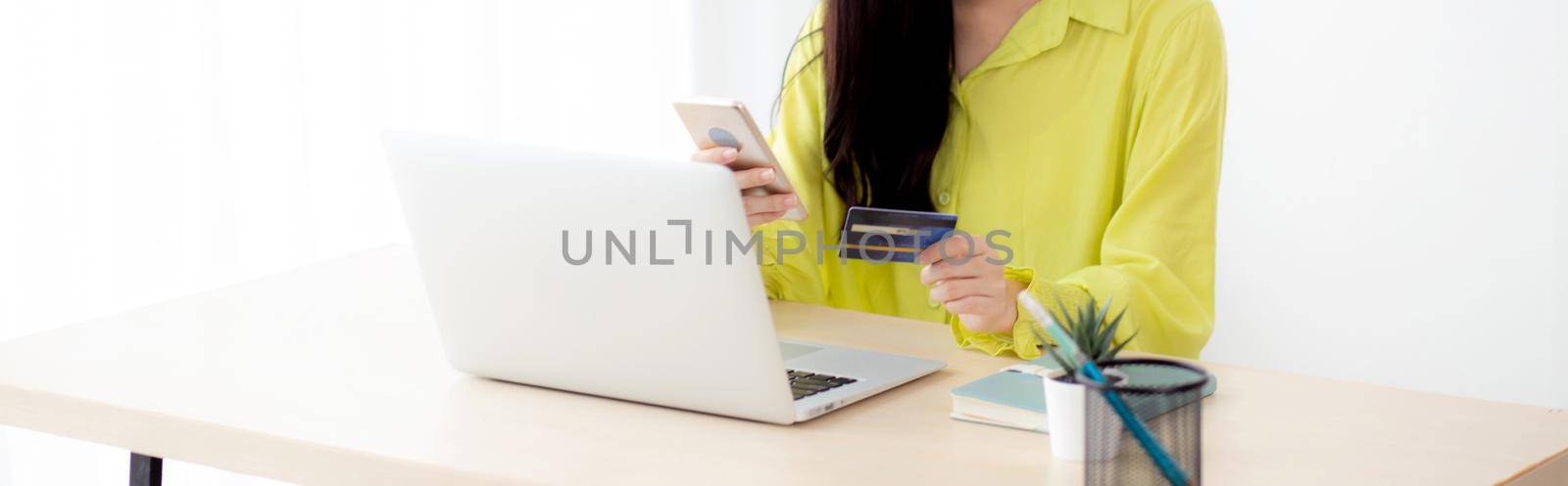 Young asian business woman using smart phone and holding credit card while online shopping and payment online with laptop computer on desk at home, female holding debit card, communication concept.