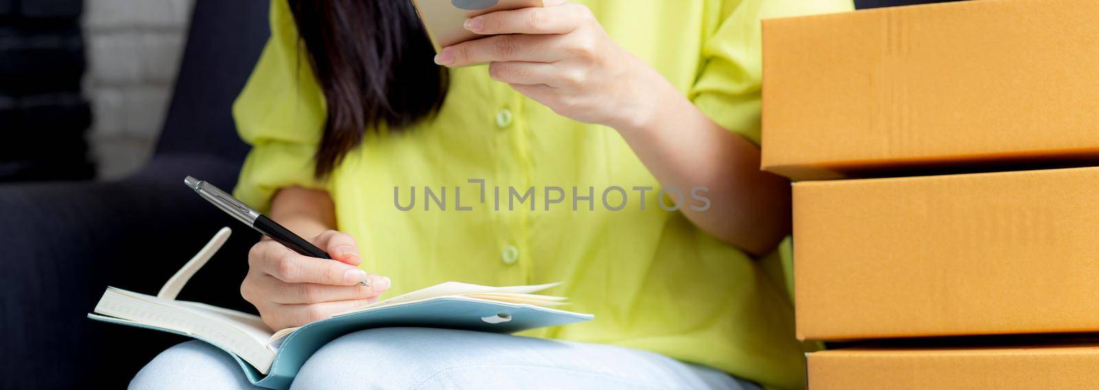 Closeup young asian woman is entrepreneur looking phone and writing order of customer with online on notebook, online shopping, logistic and delivery, female is seller working on sofa, business SME.