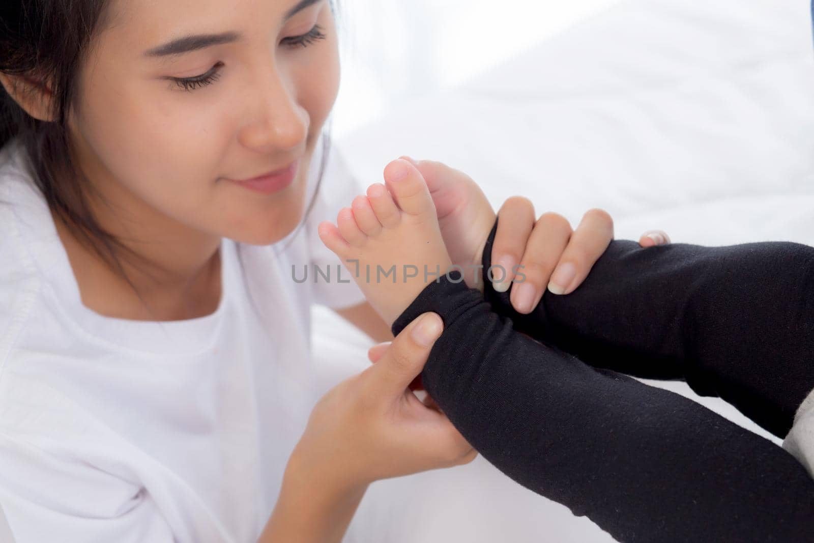 Young asian mother kiss foot of little baby girl on bed in bedroom at home, mom and newborn with relationship and bonding together, motherhood with love and care child with happy, family concept.