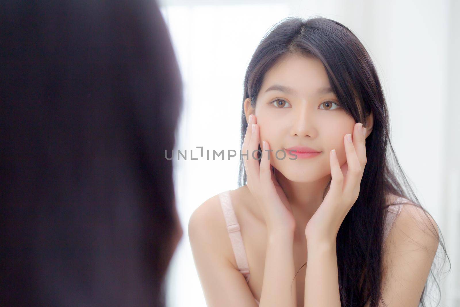 Beautiful young asian woman smiling look at mirror of checking face with skin care and cosmetic for rejuvenation and hygiene, beauty girl happy clean facial with cream or lotion and for health.