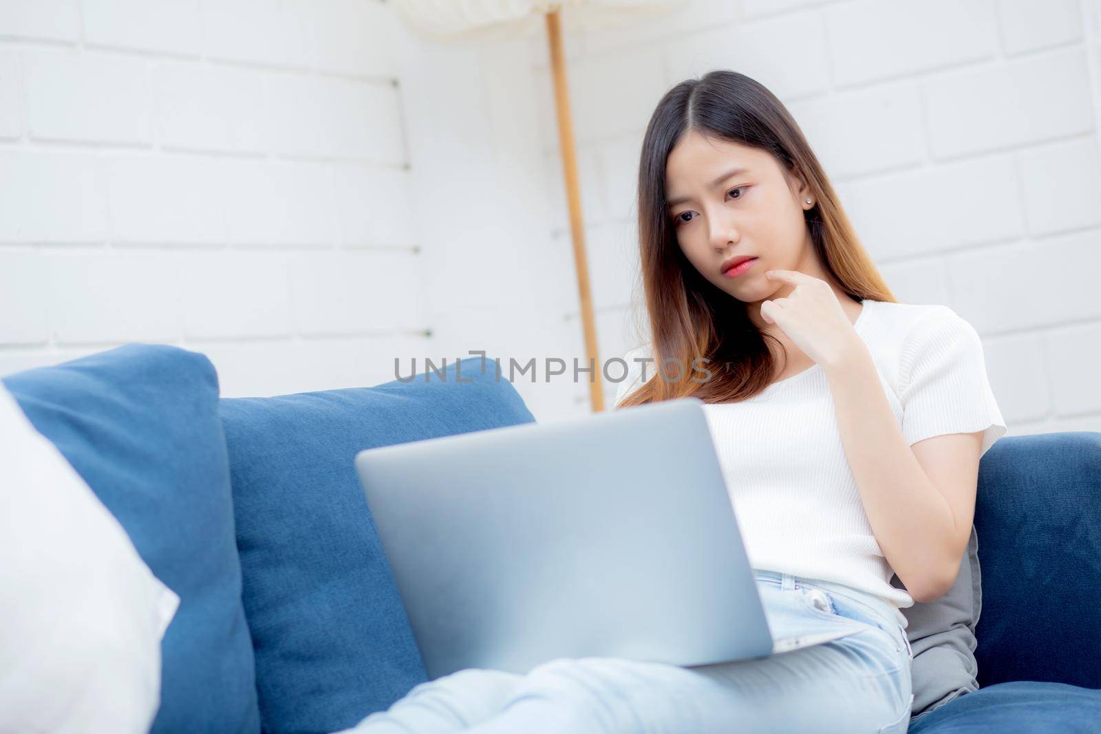 Beautiful young asian woman working on laptop computer and thinking idea on sofa at home, freelance girl sitting on couch using notebook to internet at living room, one person, lifestyle concept.