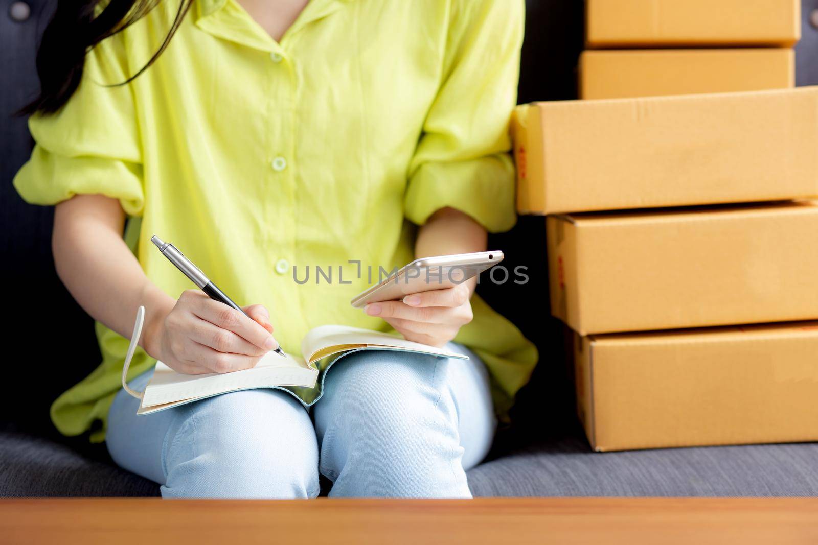 Closeup young asian woman is entrepreneur looking phone and writing order of customer with online on notebook, online shopping, logistic and delivery, female is seller working on sofa, business SME.