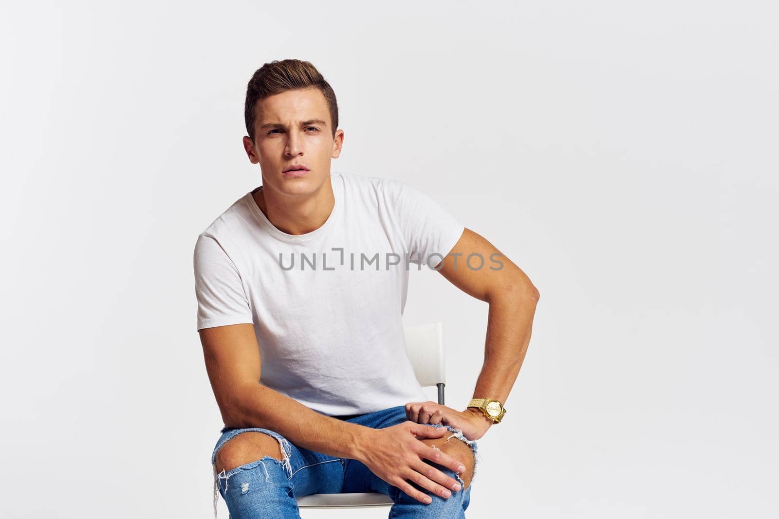 Man on a chair indoors torn jeans white t-shirt handsome face model light background. High quality photo