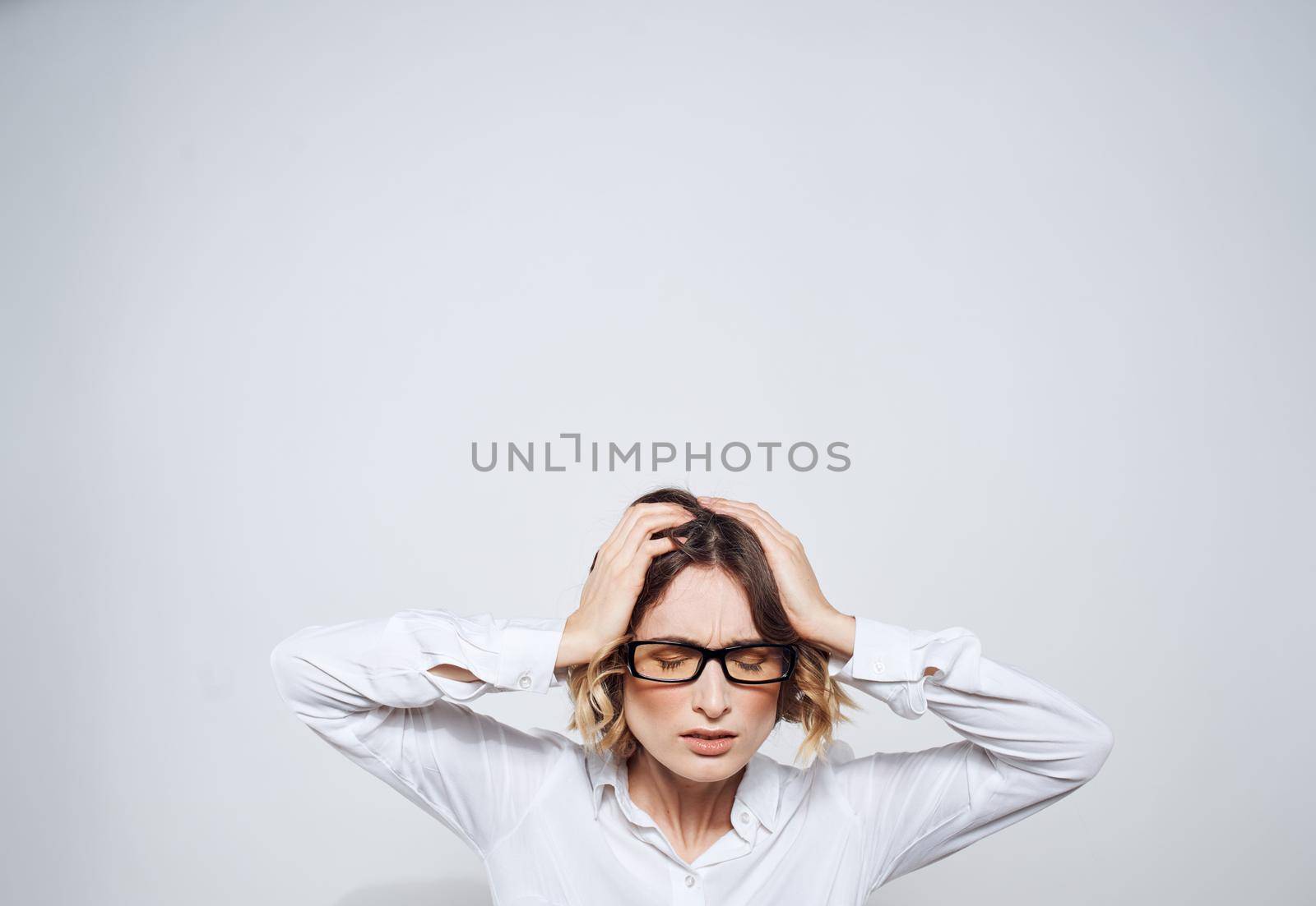 A woman in glasses and in a white shirt touches her head with her hands on a light background. High quality photo