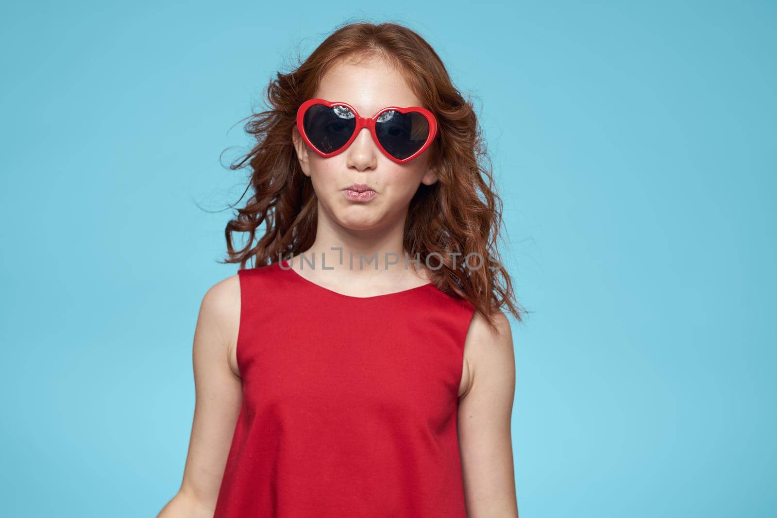 Cheerful little girl with sunglasses in the form of a heart red dress blue background. High quality photo