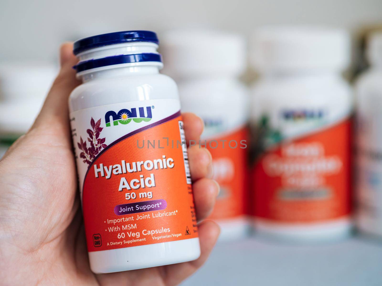 Moscow, Russia - March 09, 2021: Jar with Hyaluronic Acid capsules in female hand and others nutrition supplements in blurred background. White and orange plastic jar Hyaluronic Acid by Now Foods.