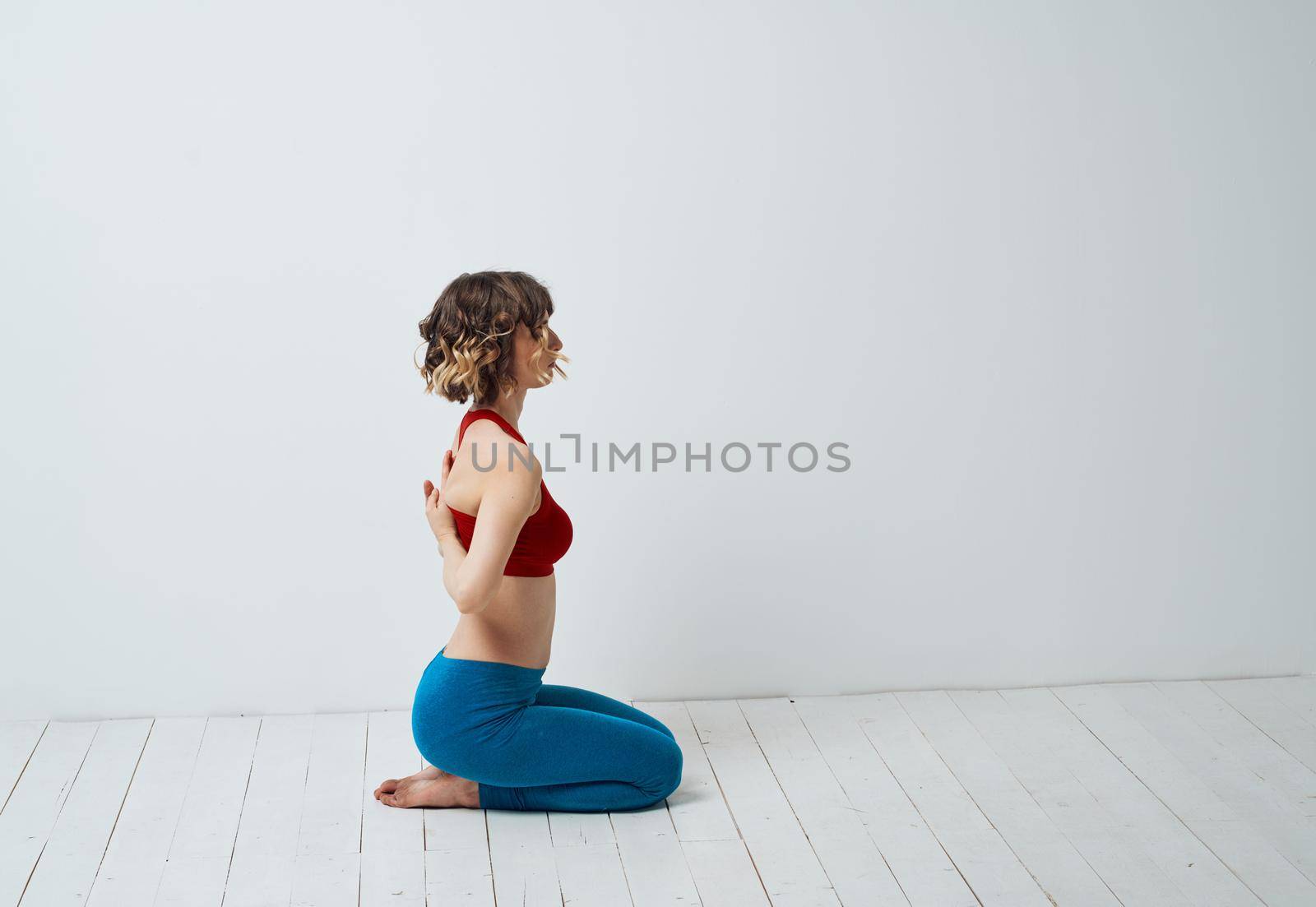 A woman in blue leggings sits on a light floor indoors and holds her hands behind her back by SHOTPRIME