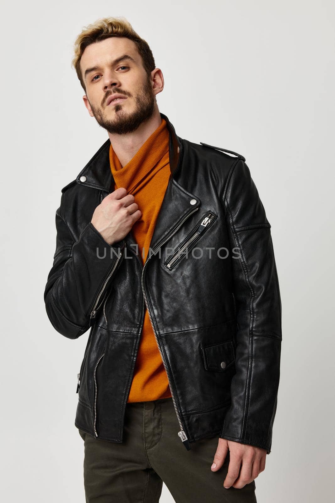 attractive young man in an orange sweater and in a leather jacket on a light background touches the collar. High quality photo