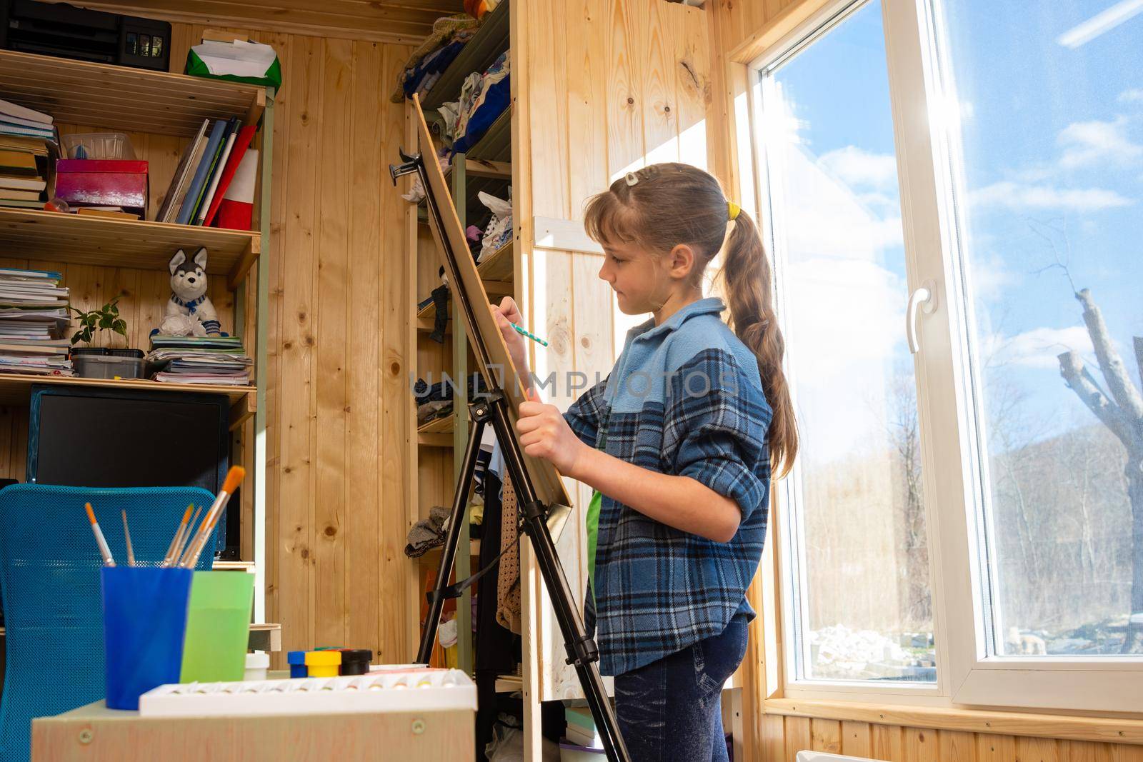 A ten-year-old girl draws on an easel by the wide window of the house
