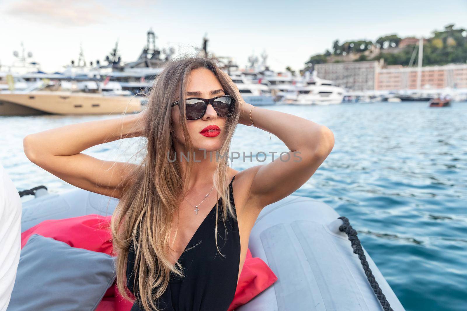 The elegant girl dressed in an evening dress of black color and sunglasses on the boat carry to the big yacht, gorgeous woman, sexual red lips, Decollete, Monaco, Monte-Carlo, is evening