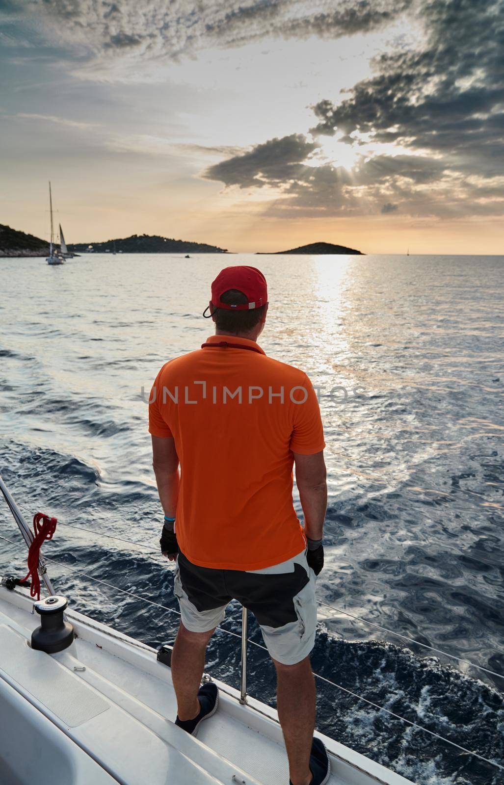 The participant of a sailing regatta is on the edge of the boat, he enjoys a victory and a sunset, he is dressing in t-shirt of orange color, sailboat and island is on background