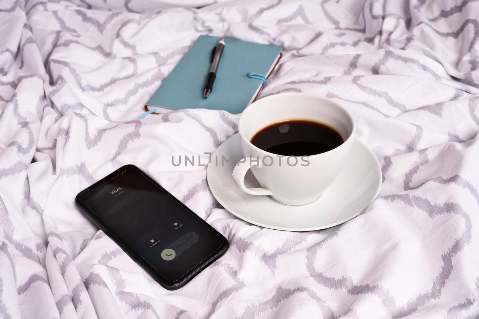 Cup of coffee on modern bed sheets, notebook pen and an incoming call from work on a mobile phone. Freelance concept. Missed calls from work.