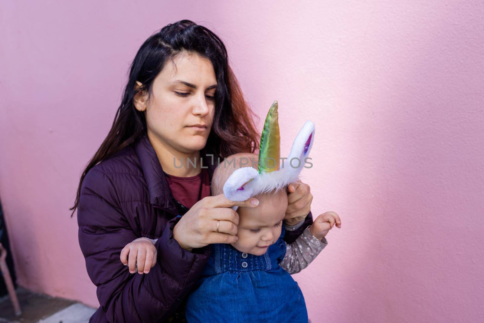 Portrait of mother in squatting position putting unicorn headband on cute baby with pink wall as background. Lovely view of happy woman holding her adorable baby girl. Happy family outdoors