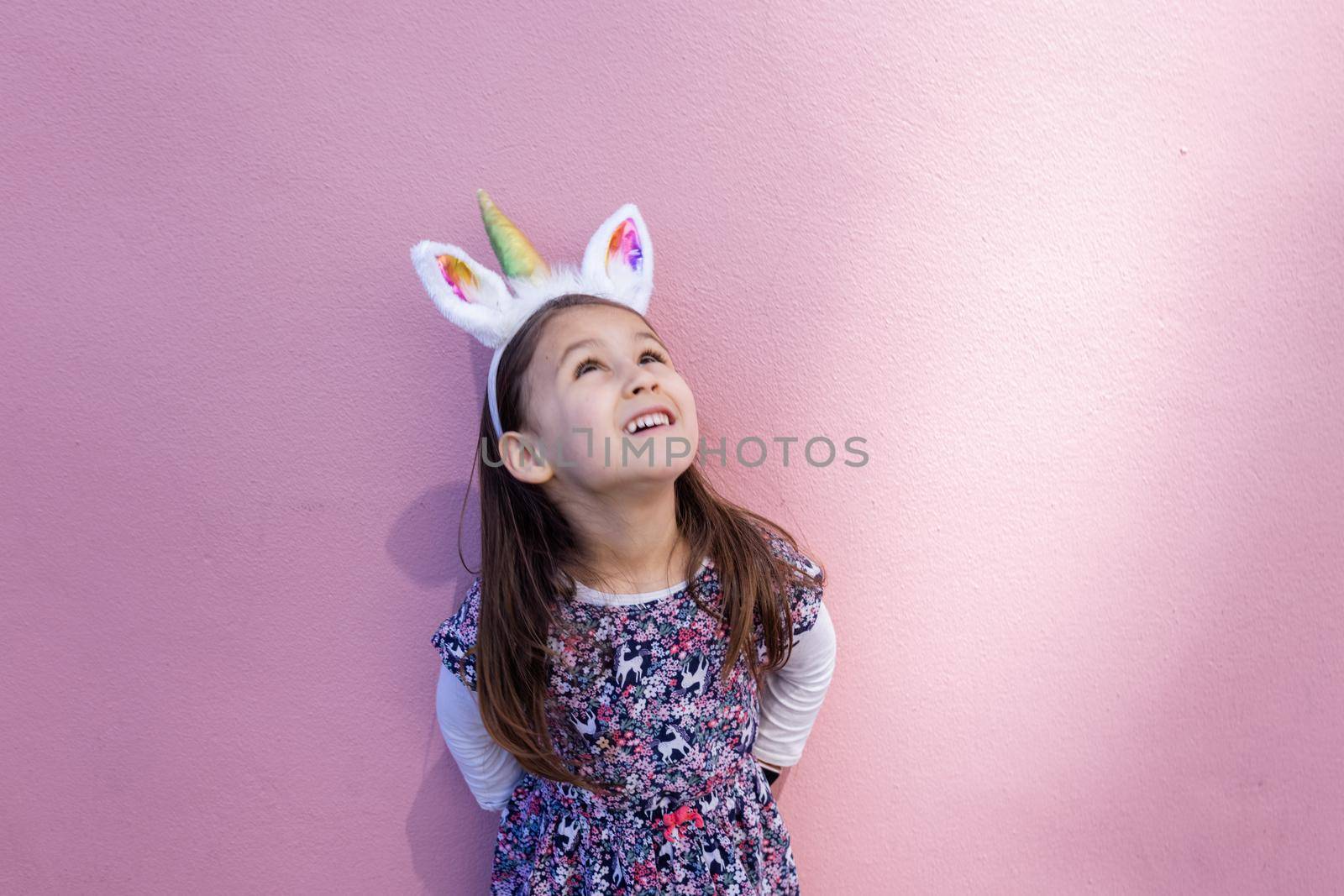 Adorable happy little girl looking up and wearing unicorn headband with pink background. Portrait of cute smiling child with unicorn horn and ears in front bright pink wall. Lovely kids in costumes