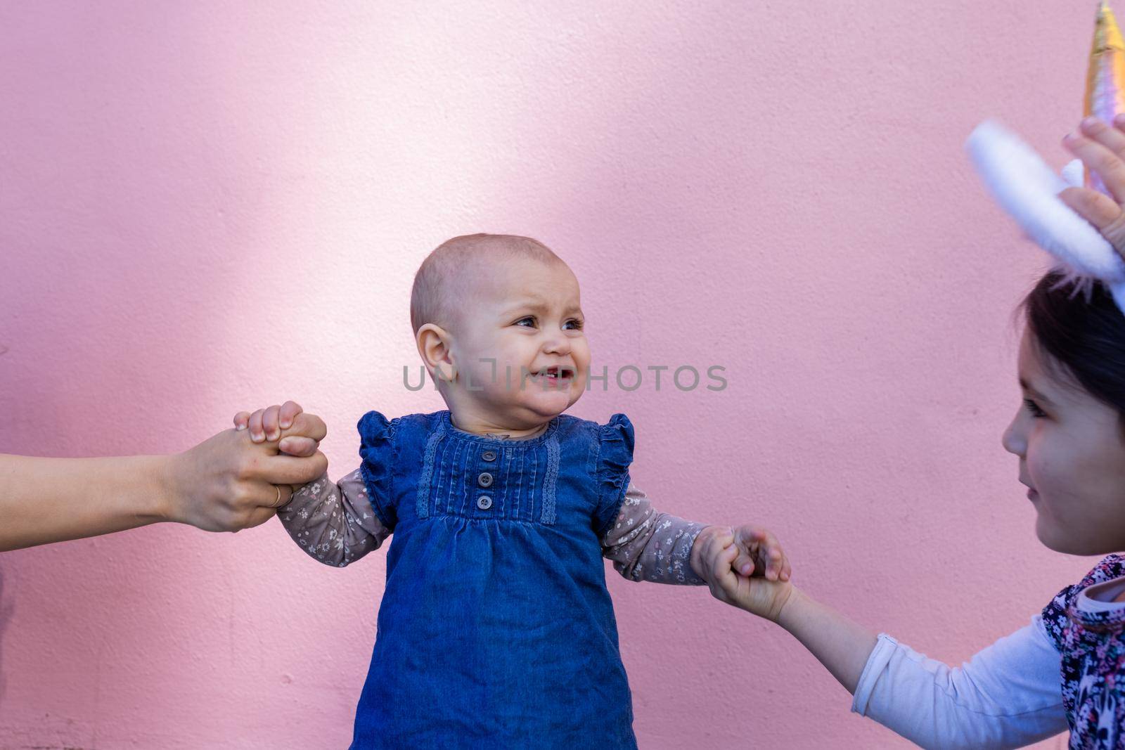 Portrait of woman and little girl holding cute smiling baby by the hands with pink wall as background. Adorable baby standing on sidewalk between her mother and older sister. Happy family outdoors