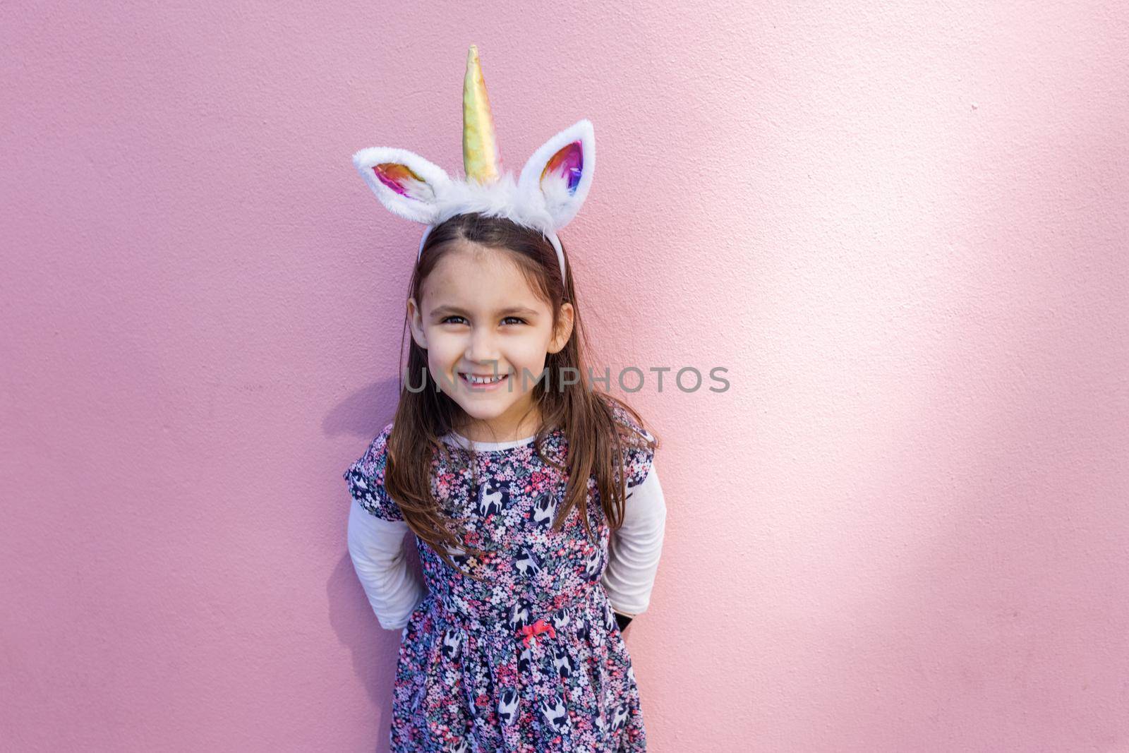 Adorable view of happy little girl wearing unicorn headband with pink background. Portrait of cute smiling child with unicorn horn and ears in front bright pink wall. Lovely kids in costumes