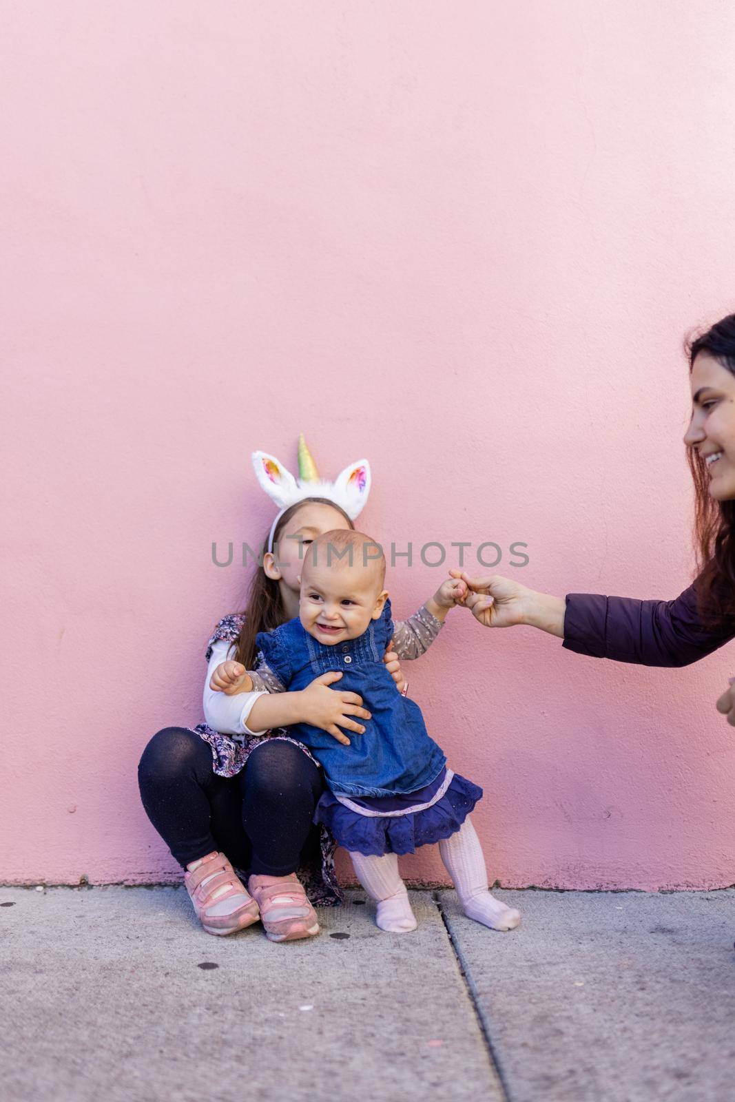 Adorable view of little girl wearing unicorn headband hugging her happy baby sister with pink background. Mother holding cute toddler hand while sitting in front bright pink wall. Lovely kids outdoors