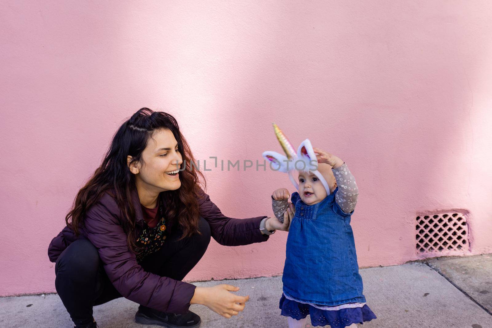 Portrait of happy mother in squatting position holding baby with unicorn headband in front of pink wall. Lovely view of smiling woman next to cute baby girl in blue dress. Happy family outdoors