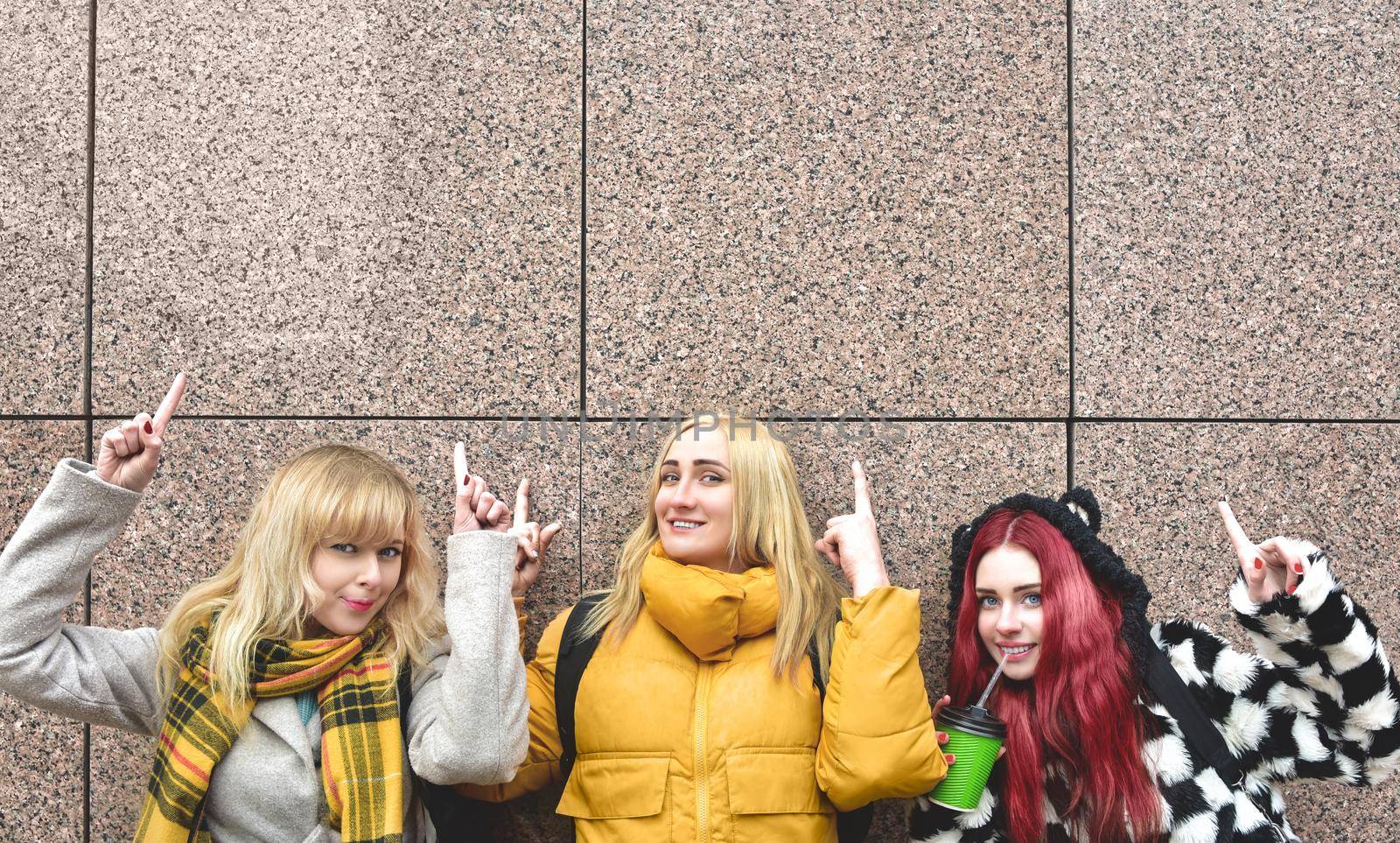 Cheerful girls at the street, show a big sale, or nice offer. Advertise concept. People, travel and tourism - women on city street show with their fingers something over gray wall background