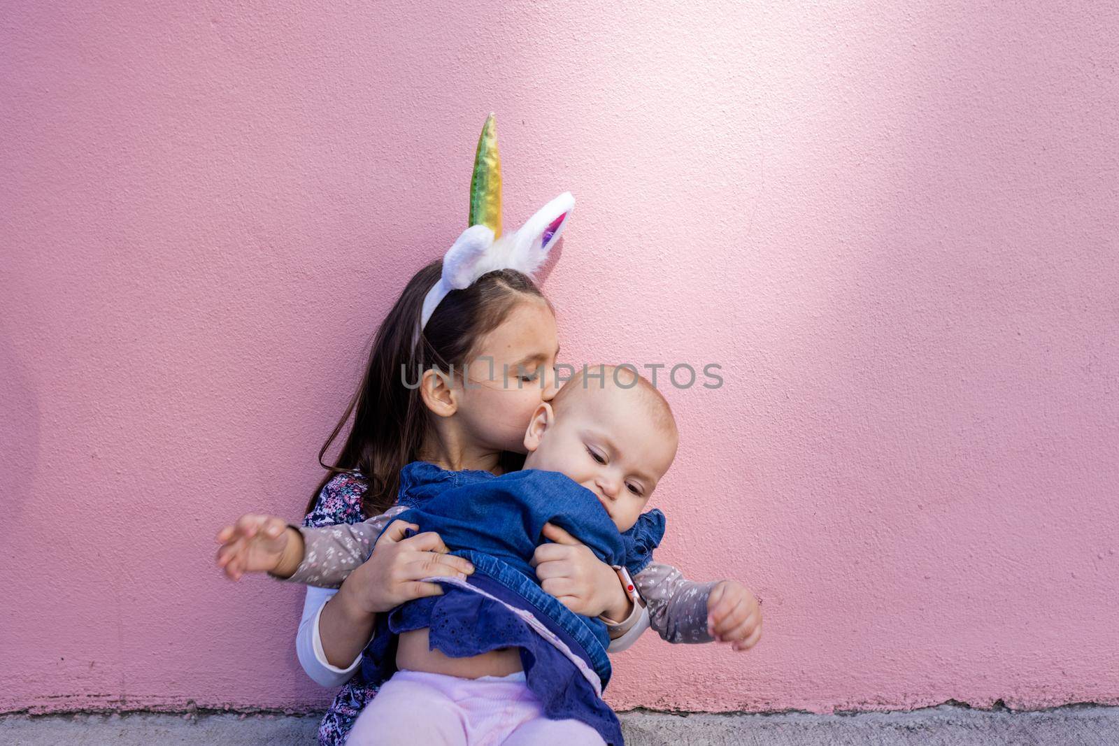 Adorable little girl wearing unicorn headband hugging and kissing a baby by Kanelbulle