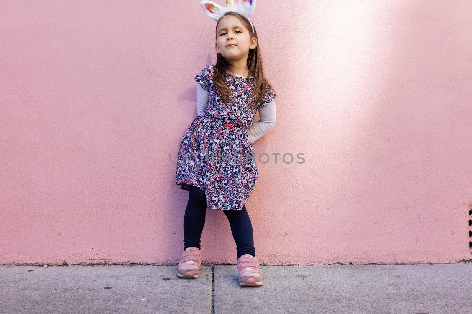 Adorable view of happy little girl wearing unicorn headband with pink wall as background. Portrait of cute smiling child with unicorn horn and ears standing on the sidewalk. Lovely kids in costumes