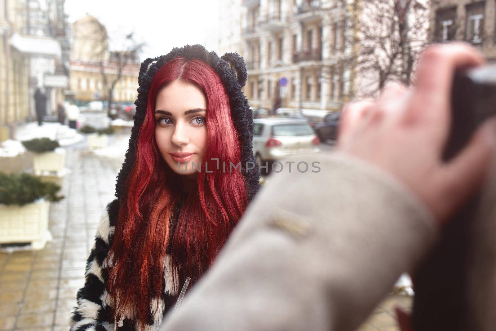 A pretty girl with red hair poses and looks at the camera while her friend takes her picture. by Nickstock