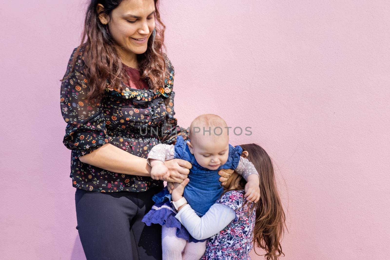 Portrait of woman helping little girl to hold cute smiling baby with pink wall as background. Adorable view of baby in arms of her older sister and mother. Happy family outdoors