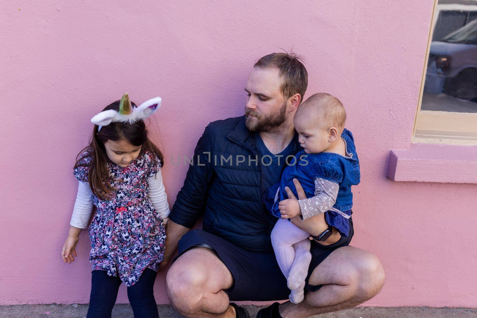 Portrait of young father with his adorable baby and older daughter in front of a pink wall. Cute child wearing unicorn headband next to baby and bearded man with pink background. Happy family outdoors