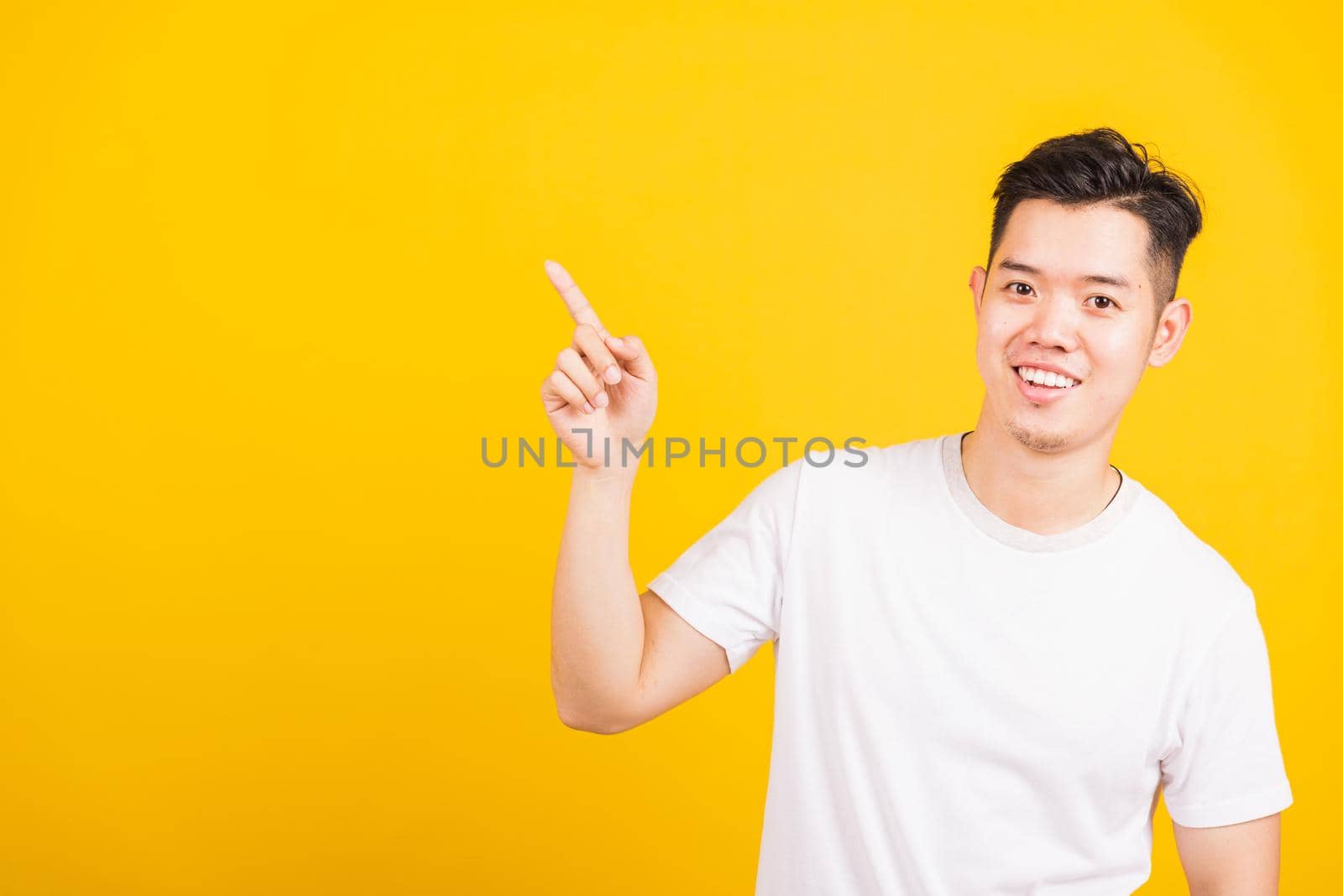 Portrait happy Asian handsome young man smiling standing wearing white t-shirt pointing finger to the side away he looking to camera, studio shot isolated yellow background