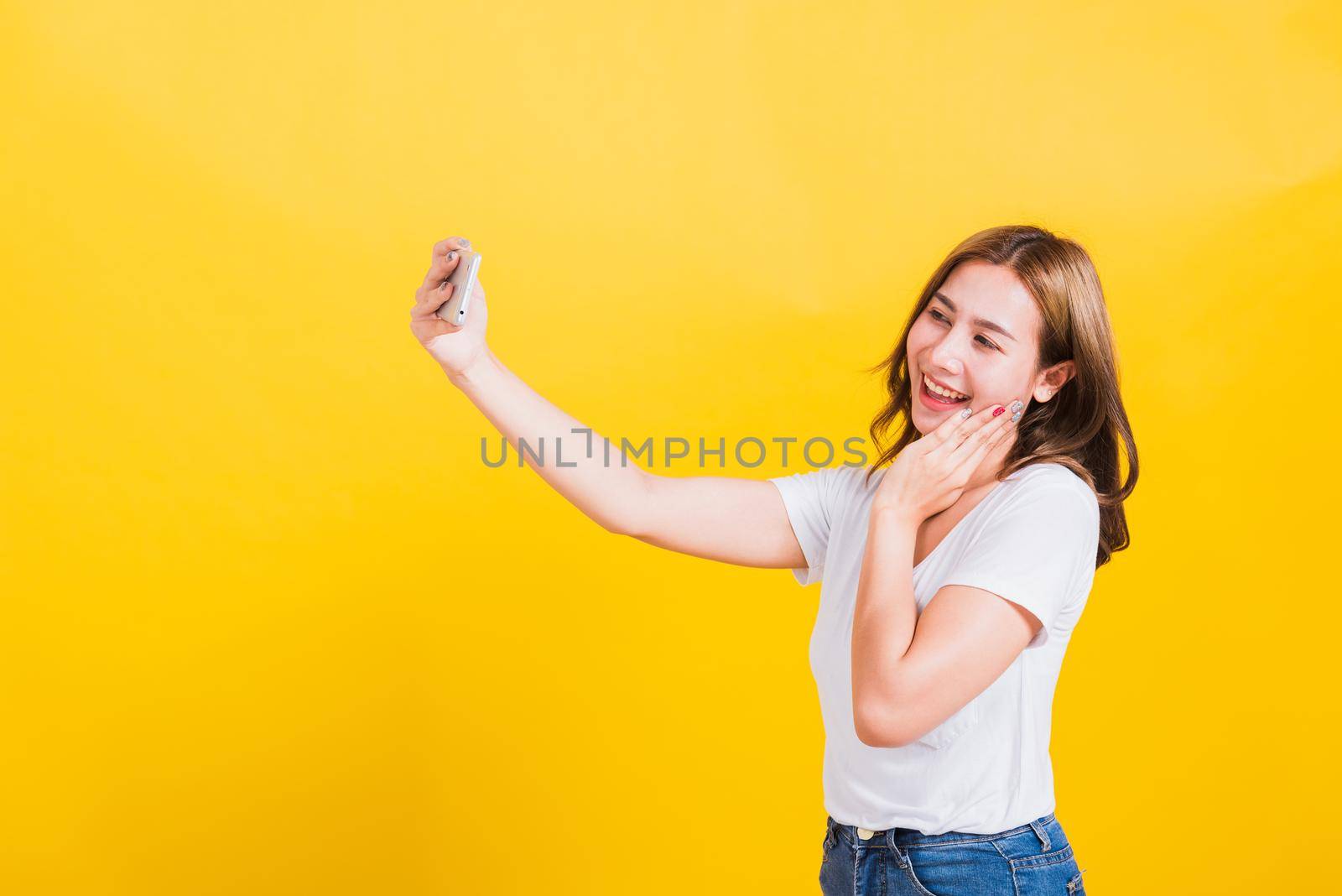 Asian Thai portrait happy beautiful cute young woman smiling wear t-shirt making selfie photo or video call on smartphone looking the phone, studio shot isolated yellow background with copy space