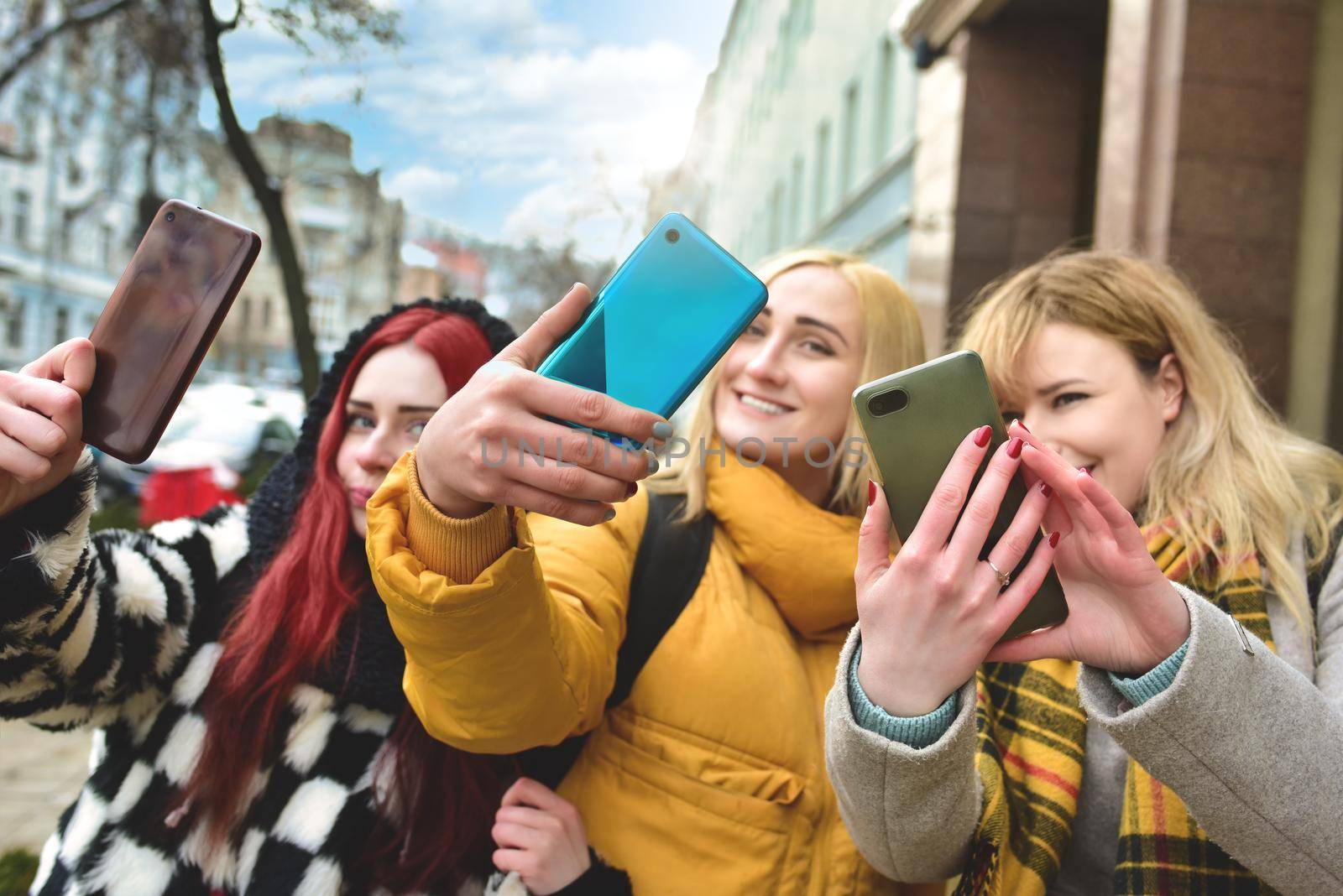 Three beautiful smiling young women walk in the city, using phone outdoors make selfies