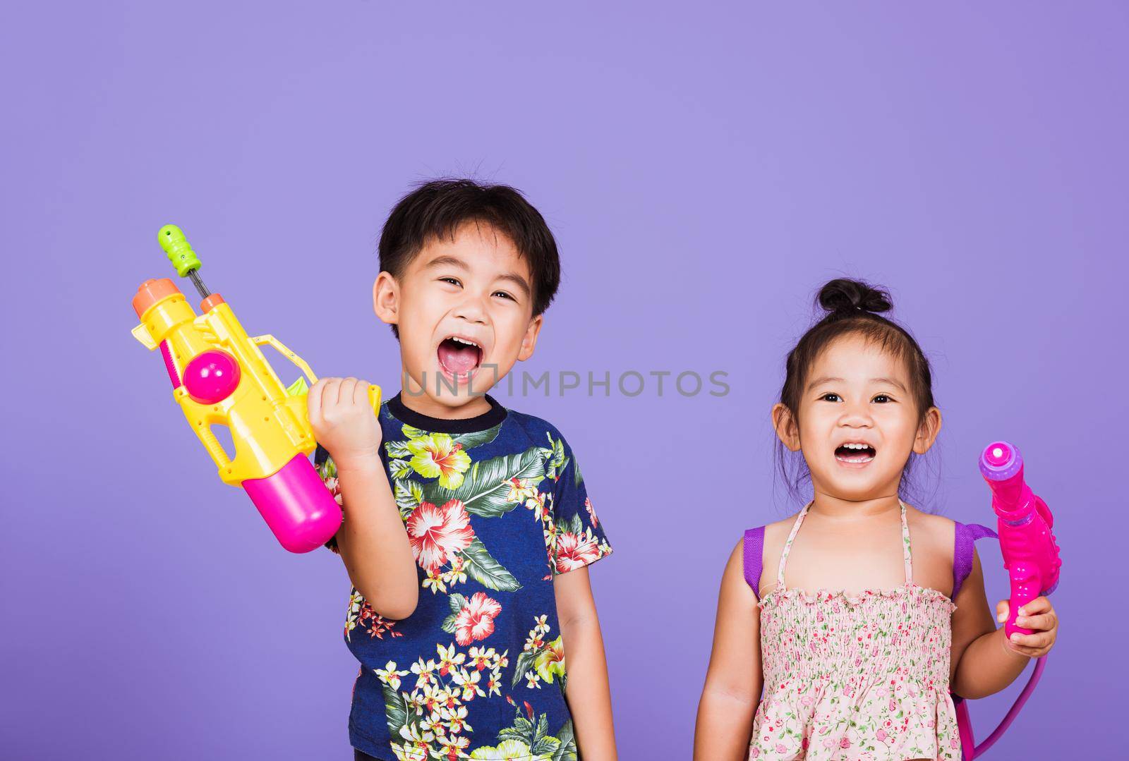 Two Happy Asian little boy and girl holding plastic water gun, Thai children funny hold toy water pistol and smile, studio shot isolated on purple background, Thailand Songkran festival day culture.