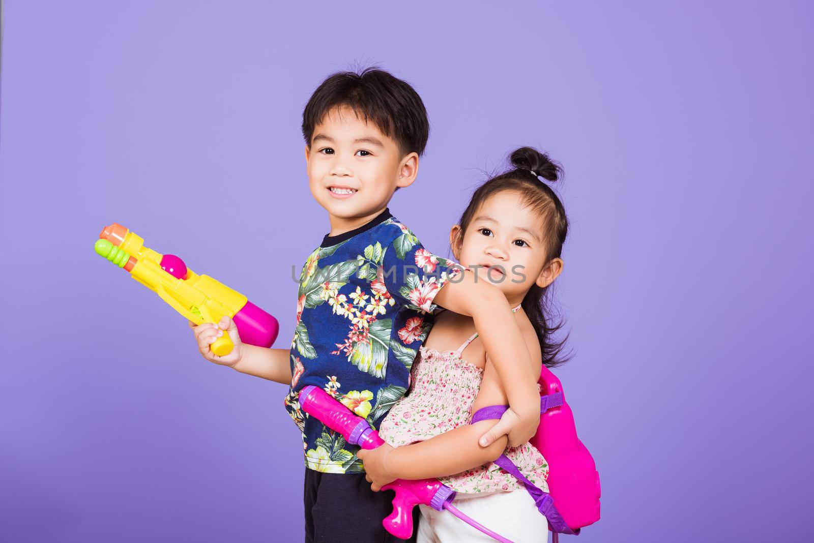 Two Happy Asian little boy and girl holding plastic water gun, Thai children funny hold toy water pistol and smile, studio shot isolated on purple background, Thailand Songkran festival day culture.